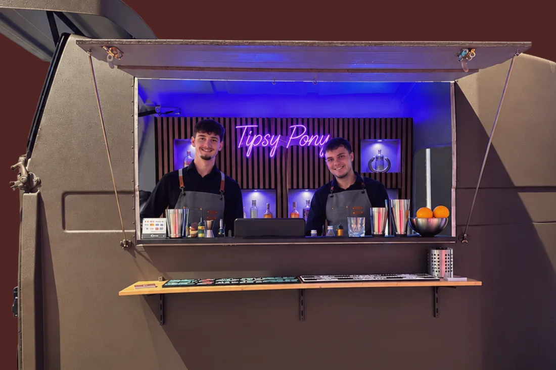 Der aufgeklappte Anhänger der Tipsy Pony Bar: zwei Barkeeper in Schürzen hinter dem Tresen, dahinter der Neonschriftzug „Tipsy Pony“, davor Shaker und eine Schale mit Orangen.